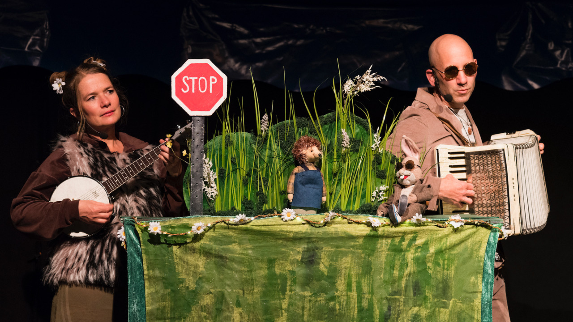 Zwei Schauspielende machen Musik. Davor steht ein Tisch mit aufgebautem Bühnenbild: Die Puppen von Hase und Igel sitzen am Straßenrand.